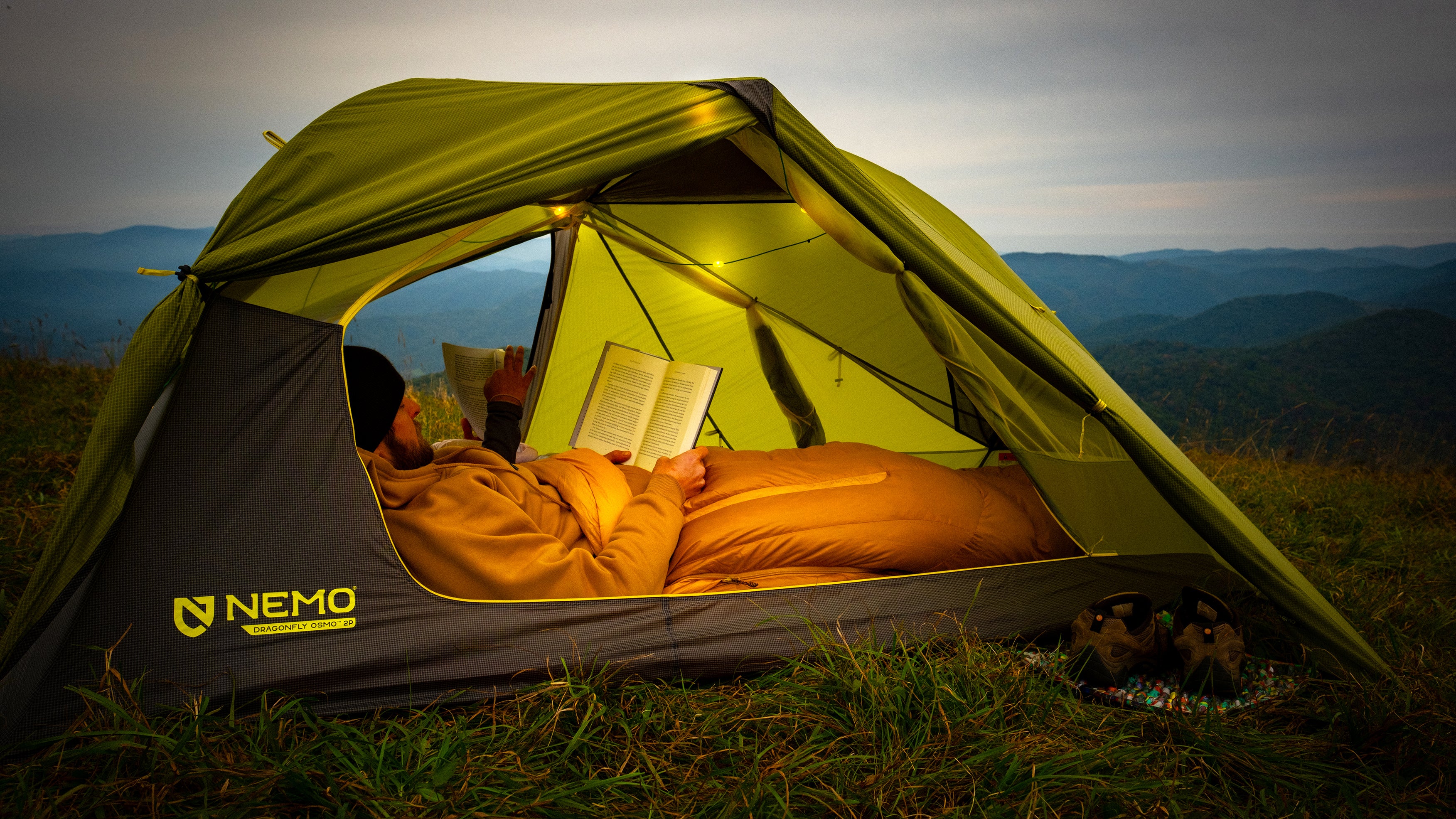 Person lying in a green tent, reading a book, and wrapped in a yellow sleeping bag with the brand logo 